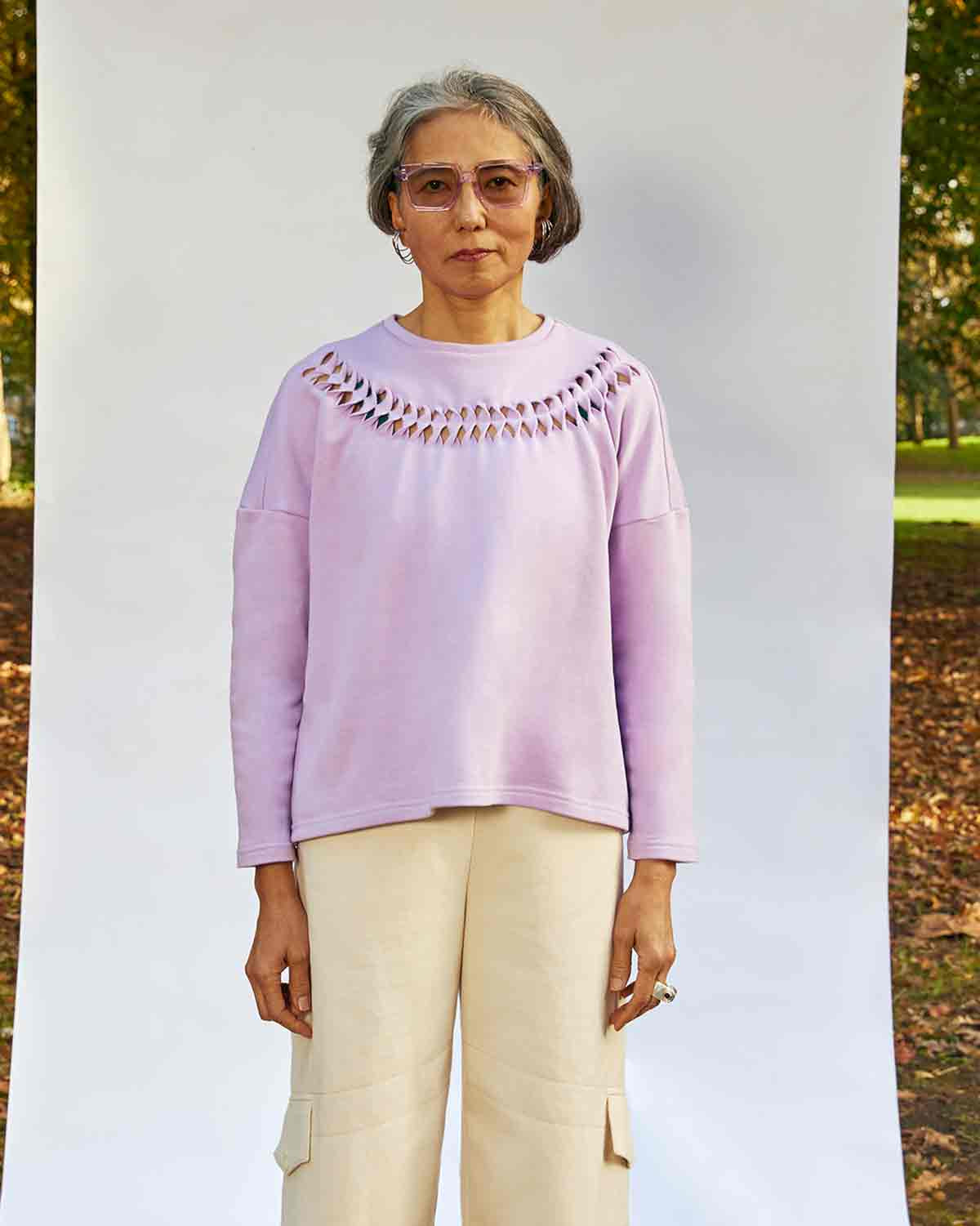 Women standing face on wearing the Anni Sweatshirt in Lilac with detailing where the fabric is cut, twisted and stitched back in place. She is standing in front of a white background in a park