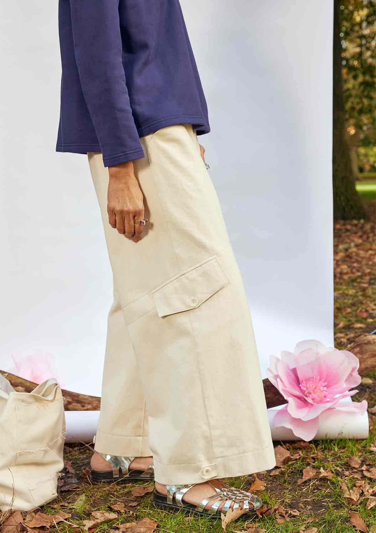 Woman standing side on wearing the Asmuss Eddy Cargo Trousers and Asmuss Anni Sweatshirt in Navy. The hem buttons are loose. She is standing in front of a white background, weighted down by a branch in a park with an Asmuss Tote Bag and pink paper flower on the ground behind her