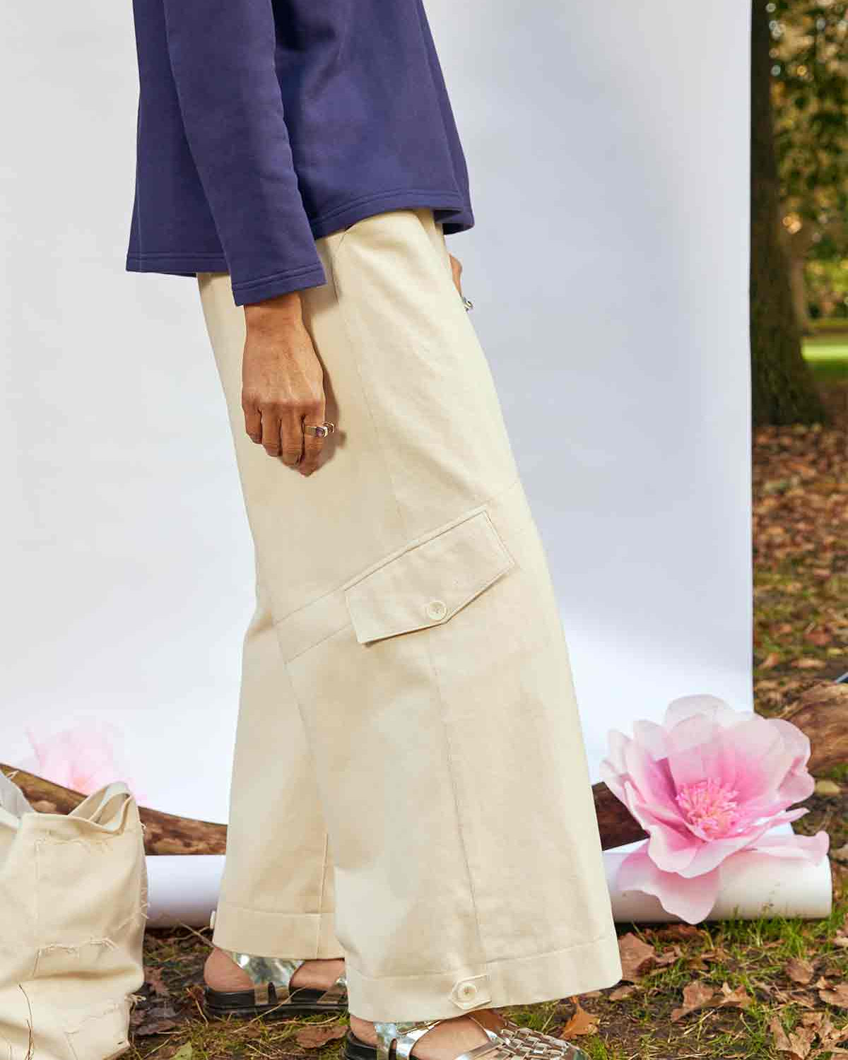 Woman standing side on wearing the Asmuss Eddy Cargo Trousers and Asmuss Anni Sweatshirt in Navy. The hem buttons are loose. She is standing in front of a white background, weighted down by a branch in a park with an Asmuss Tote Bag and pink paper flower on the ground behind her