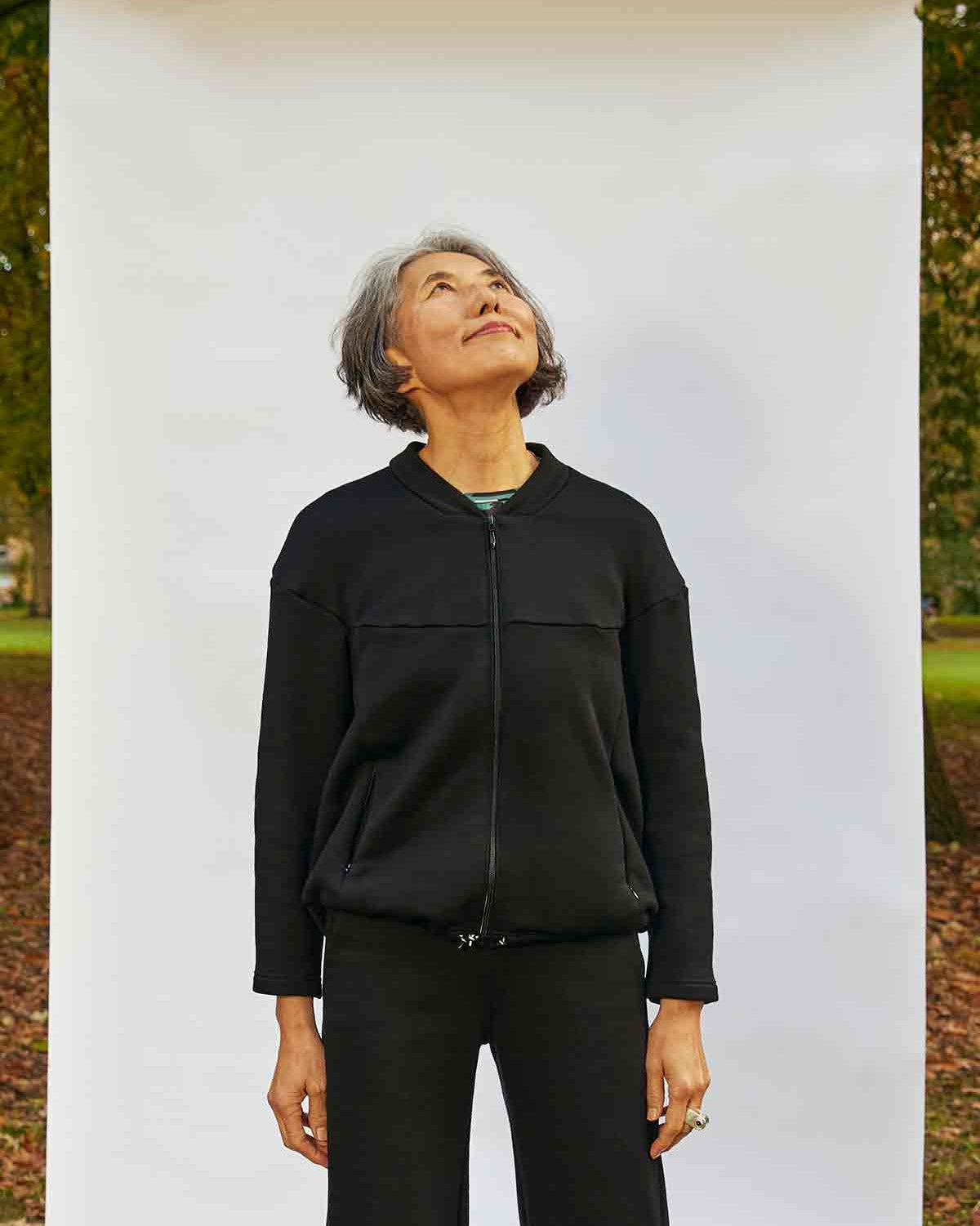 Woman, looking up, standing with her hands by her sides. She is wearing the Asmuss Hepworth Zip Through Sweatshirt zipped up.  She is standing in front of a white background outside in a park