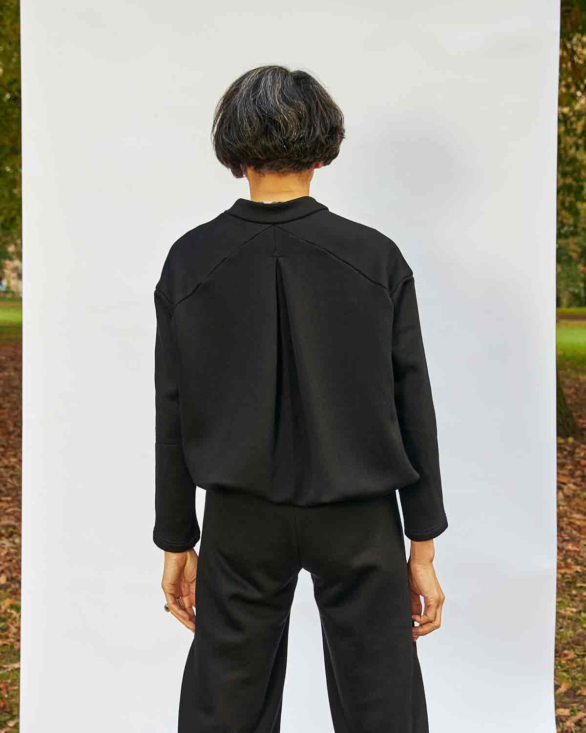 Woman stadning back on showing the inverted pleat detail on the Asmuss Hepworth Zip Through Sweatshirt.  She is standing in front of a white background that is outside in a park