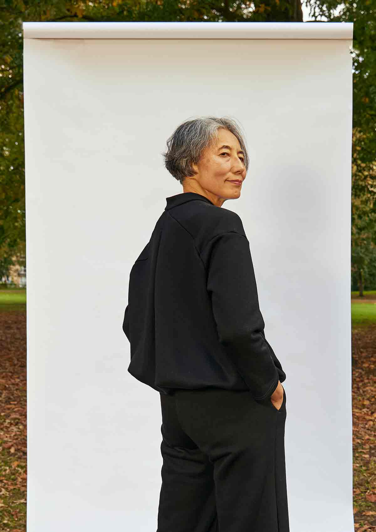 Woman, standing side on. She is wearing the Asmuss Hepworth Zip Through Sweatshirt and can see the volume and shape created by the inverted pleat back detail and drawstring in the hem.  She is standing in front of a white background outside in a park 