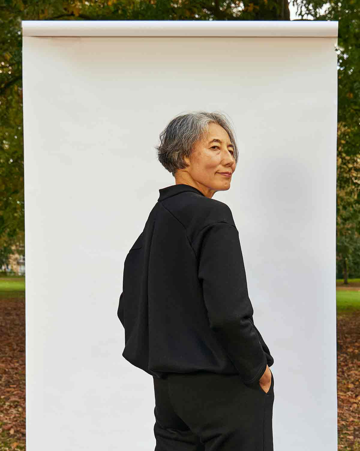Woman, standing side on. She is wearing the Asmuss Hepworth Zip Through Sweatshirt and can see the volume and shape created by the inverted pleat back detail and drawstring in the hem.  She is standing in front of a white background outside in a park