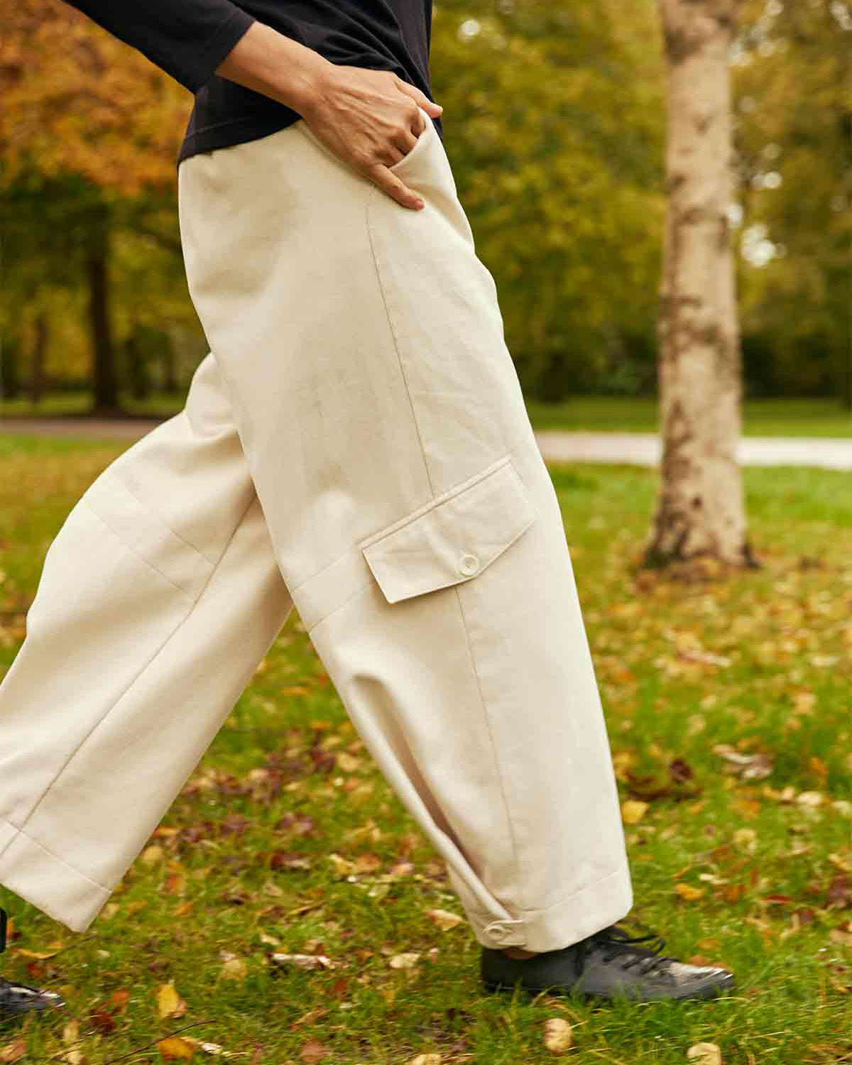 Woman walking in a park with her right leg forward showing the tab pocket detail on the leg and the buttoned hem of the ercu Asmuss Eddy Cargo Trousers.  Her hand is in the hip pocket.