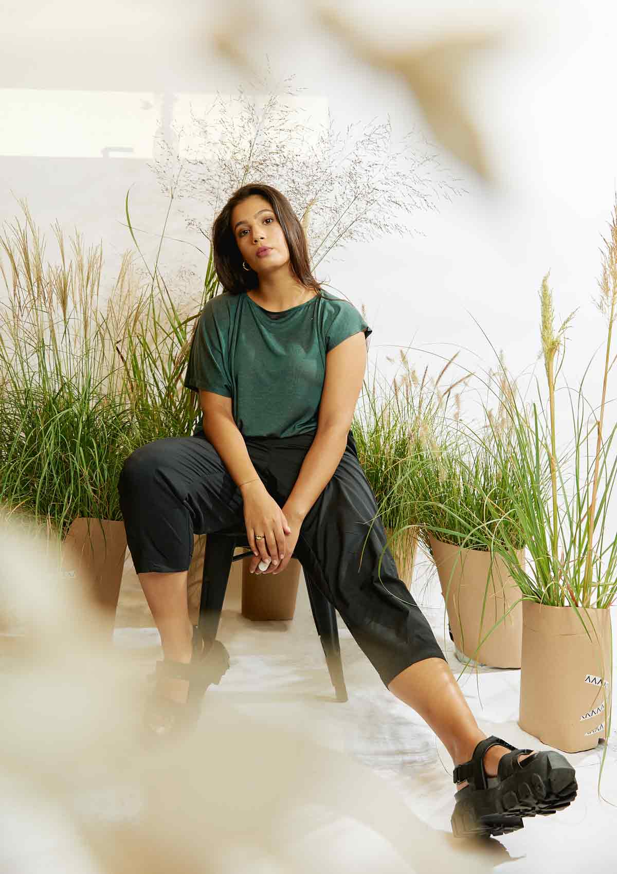 Woman sitting amongst grasses with one leg stretched out wearing the pine green Heed T-shirt and the black Asmuss Oval Trousers