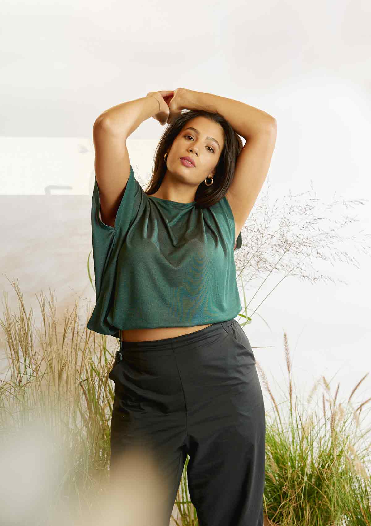 Woman standing amongst grasses the pine green Heed T-shirt that has had its drawstring tightened to create a cropped style