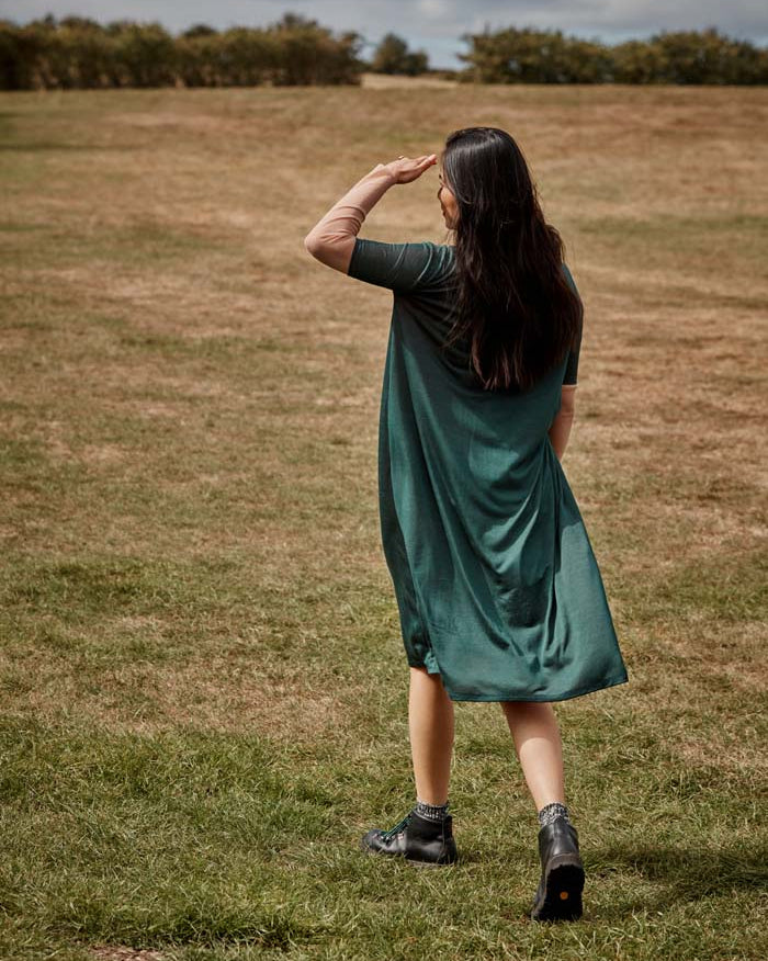 Woman walking away wearing the fluid moving Asmuss Asymmetric Pleat Dress in Pine Green. Slim arms, loose fitting body with pleat detail and pocket it makes a great dress for your everyday adventures. Made from a blend of recycled polyester and Tencel a great sustainable fabric. A stylish, sustainable addition to the modern woman's wardrobe.