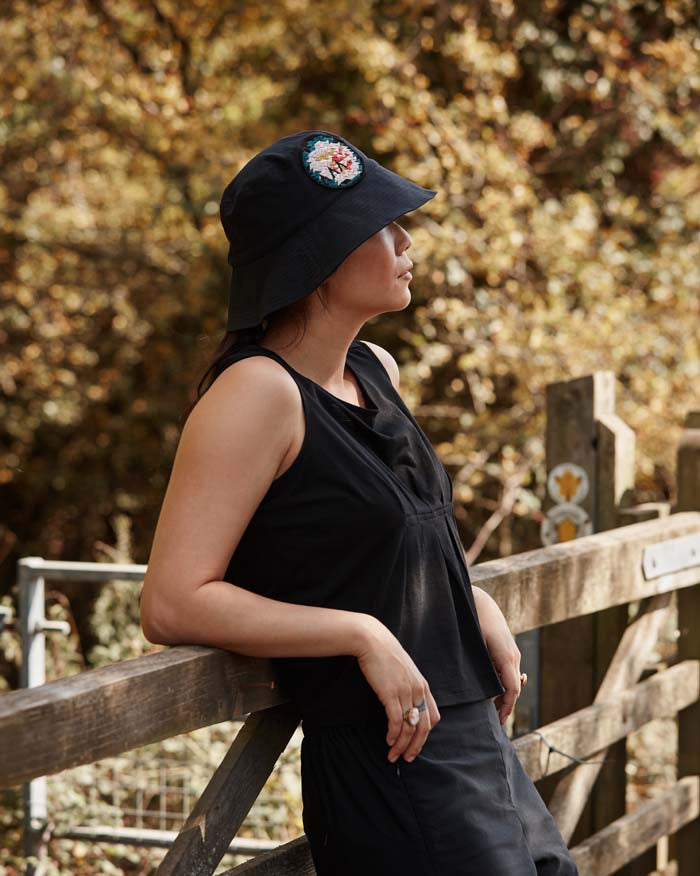 Woman leading on her elbows relaxing during a walk in the countryside wearing the Asmuss Wide Brim Bucket Hat in black with rose embroidery badge. The perfect sun hat to keep the sun off your face but still look good and with water resistance to keep the rain off too.