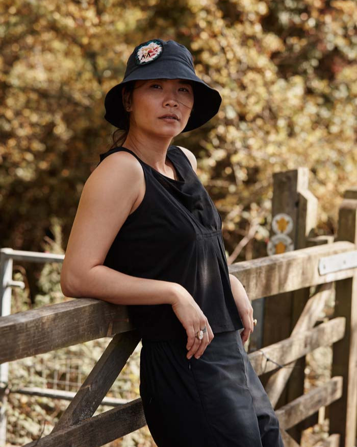Woman leaning on a wooden gate wearing the Asmuss Pleated Tank in Black. Made in small batches in the UK from a luxury wool blended with temperature regulating 37.5 Technology. A perfect summer elevated essential for all your everyday adventures.