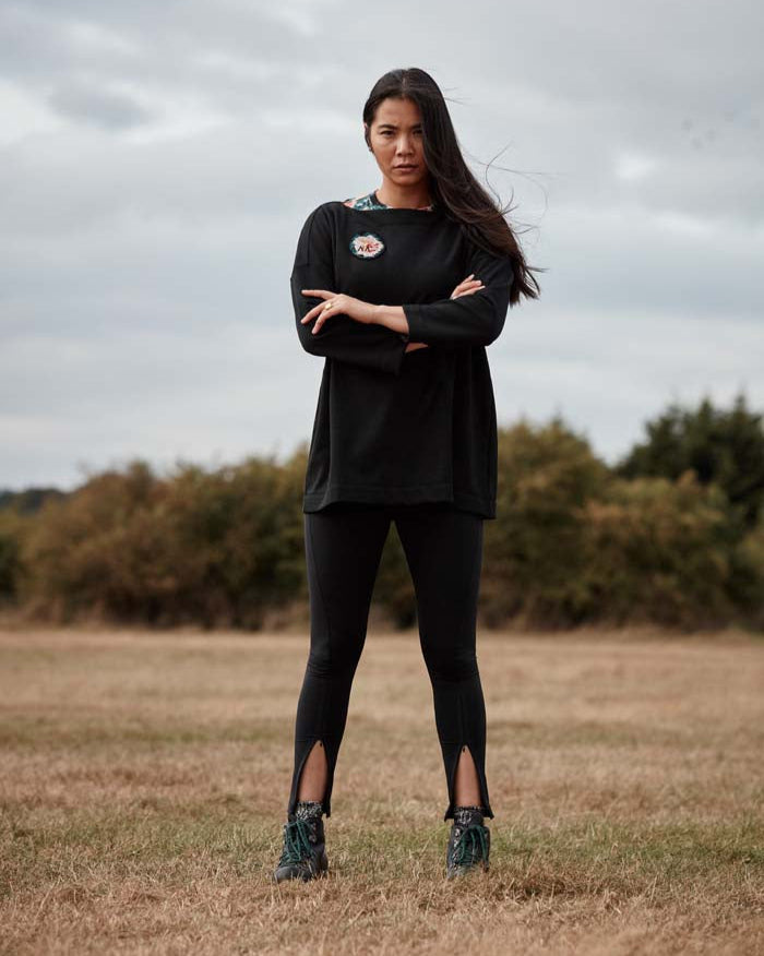 Woman standing with arms crossed in a fieldwearing the Asmuss Twisted Leggings with zip detail on lower front leg, pocket in front waistband and drawstring to keep them snug. Made in the UK from recycled nylon and elastane for 4 way stretch. Sustainable activewear that looks great on for all your everyday adventures including lazing on the couch or out running.