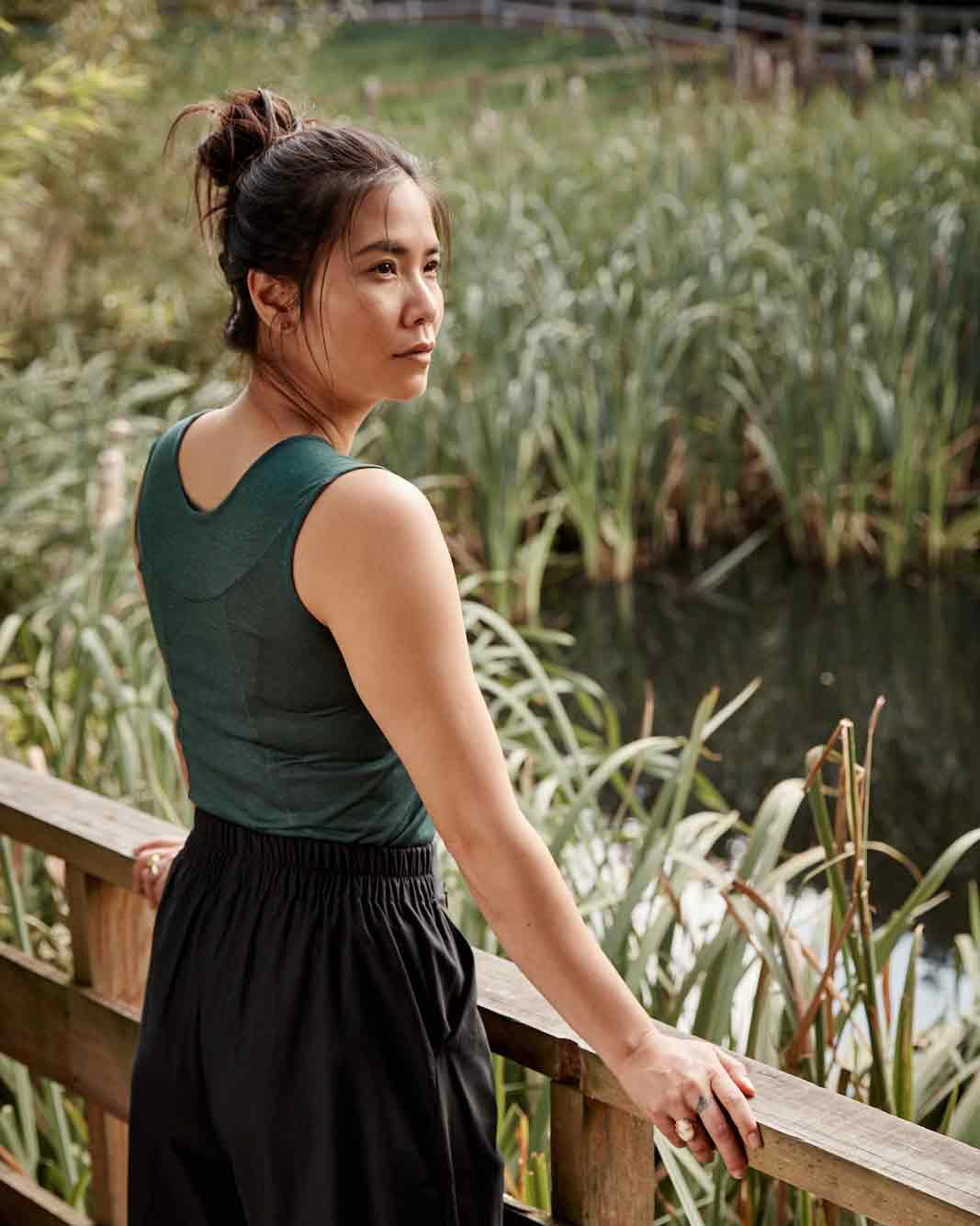 Back view of woman wearing the Asmuss Panelled Tank in Pine Green leaning on a railing near a pond filled with reeds. Stylish and sustainable lifestyle womenswear for your everyday adventures and activewear.