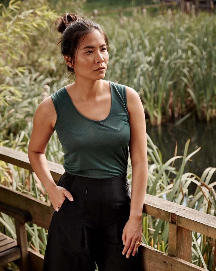 Woman wearing the Asmuss Panelled Tank in Pine Green leaning on a railing near a pond filled with reeds. Stylish and sustainable lifestyle womenswear for your everyday adventures and activewear.