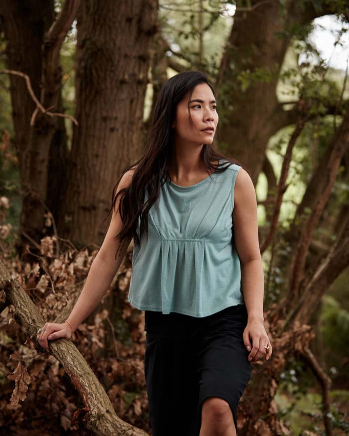 Woman wearing the Asmuss Pleated Tank in sea green while exploring a forest. Made in the UK from a blend of wool and 37.5 technology to help keep your ideal body temperature so it is perfect for travelling with or wearing at home or office. The pleat detail and darts to prevent gaping make it a great elevated essential for all your everyday adventures.