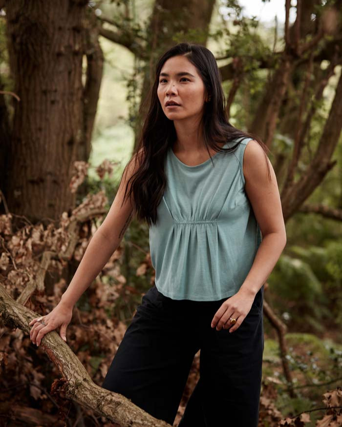 Woman wearing the Asmuss Pleated Tank in sea green while exploring a forest. Made in the UK from a blend of wool and 37.5 technology to help keep your ideal body temperature so it is perfect for travelling with or wearing at home or office. The pleat detail and darts to prevent gaping make it a great elevated essential for all your everyday adventures.