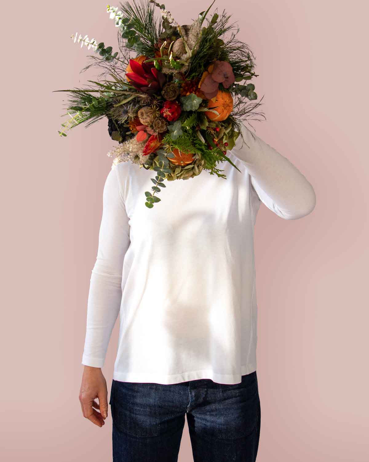 A person wearing a white long sleeve t-shirt with a crew neckline, featuring an inverted pleat in the back, standing with a bouquet of flowers in front of their face.