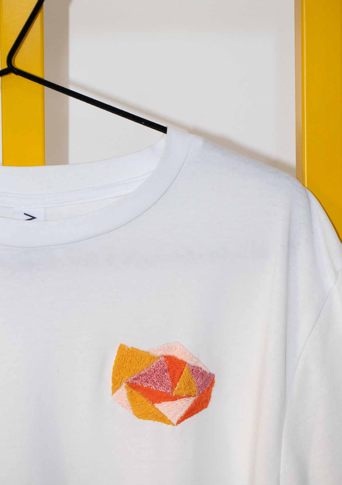 close up of white Evelyn tee-shirt showing hand embroidery of a geometric version of a rose in pinks, rust and mustard, placed on chest at left hand side of tee-shirt. 