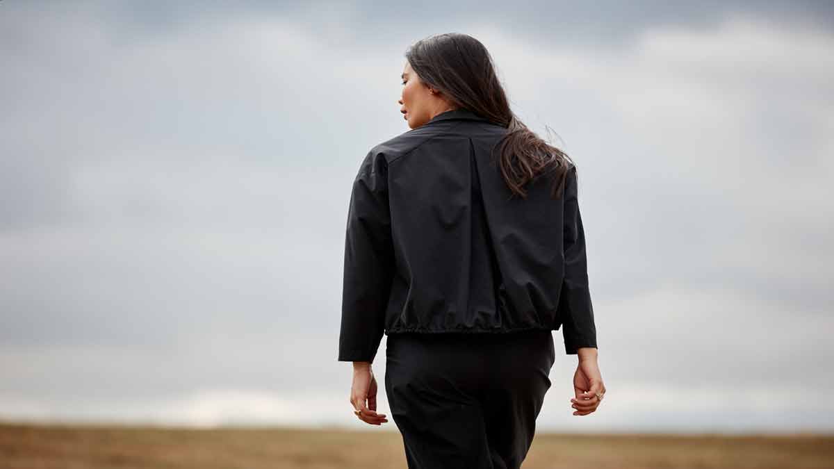 Woman walking away from the camera towards the horizon while looking back over her shoulder. She has long black hair and is wearing the Asmuss Roam Jacket in black