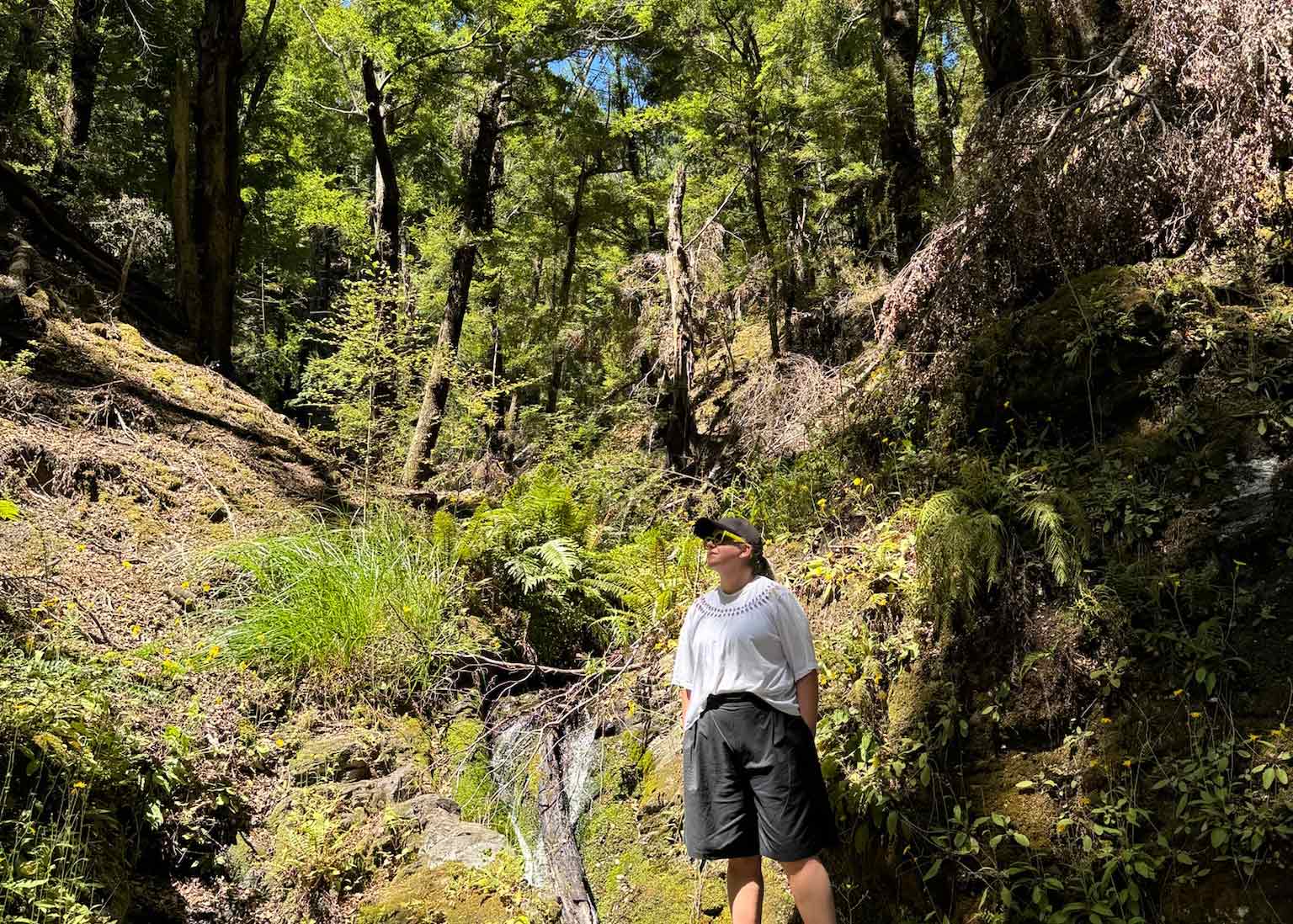 Asmuss designer and cofounder Clare standing beside a rocky stream surrounded by beech forest on the Fernburn track near Wanaka New Zealand