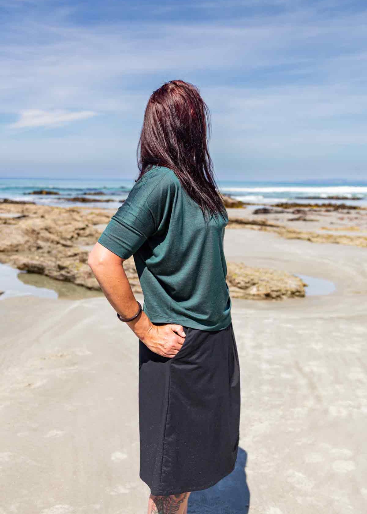 Woman standing side on in front of rock pools on a sandy beach wearing the  green Vivienne Cowl Neck Top showing the sleeve and drape of the top