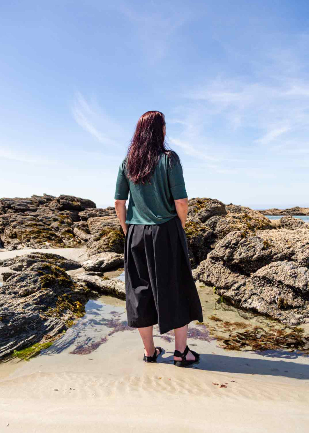 Woman standing in front of rock pools on a sandy beach wearing the black Kate Culottes and green Vivienne Cowl Neck Top showing the pleat details on the back of the culottes