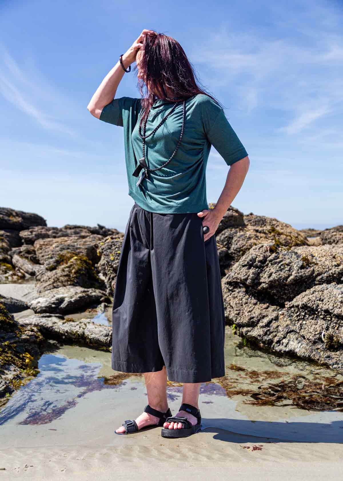 Woman standing in front of rock pools on a sandy beach, with her hand in her hair, wearing the black Kate Culottes and green Vivienne Cowl Neck Top
