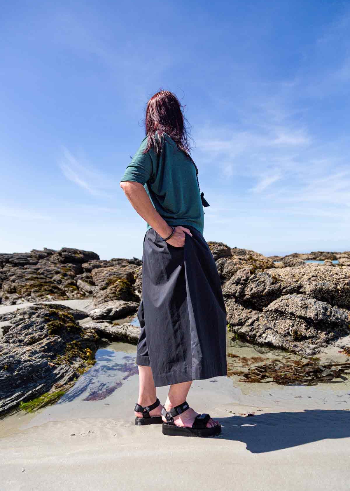 Woman standing side on in front of rock pools on a sandy beach wearing the black Kate Culottes and green Vivienne Cowl Neck Top. She has her hand in one of the pocket sand shows the side pleat in the culottes