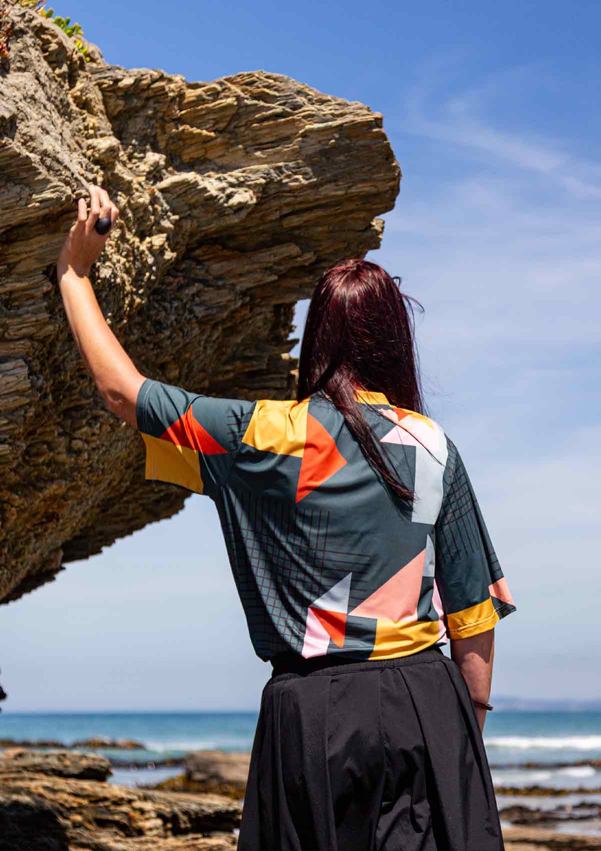 Woman standing looking toward the blue sea with her left hand leaning on rocks the are overhead. She is wearing the Asmuss Drew T-shirt