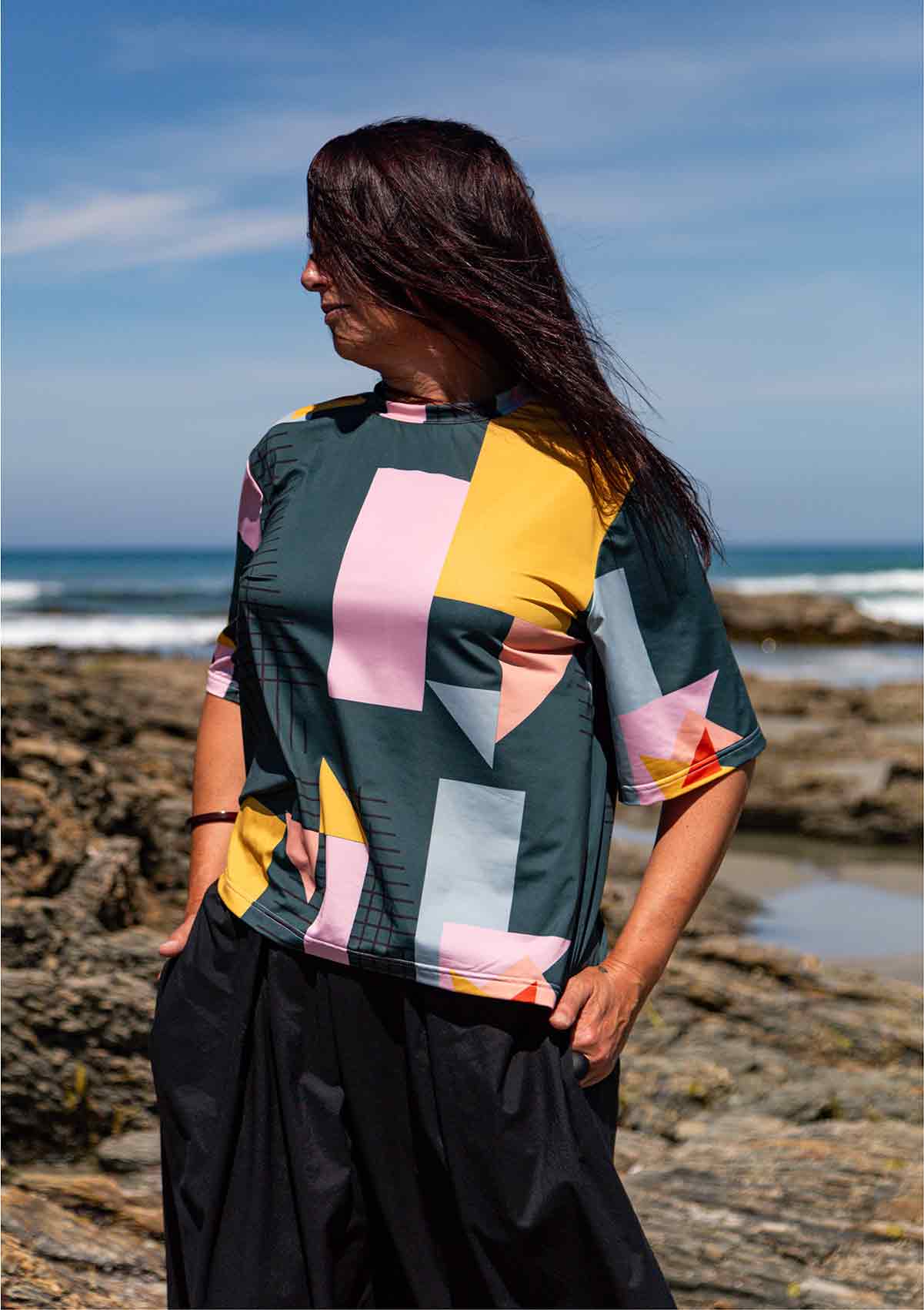 Woman wearing the Asmuss Drew T-shirt. She has long dark hair, is looking right and is standing on rocks in front of the blue sea and blue sky