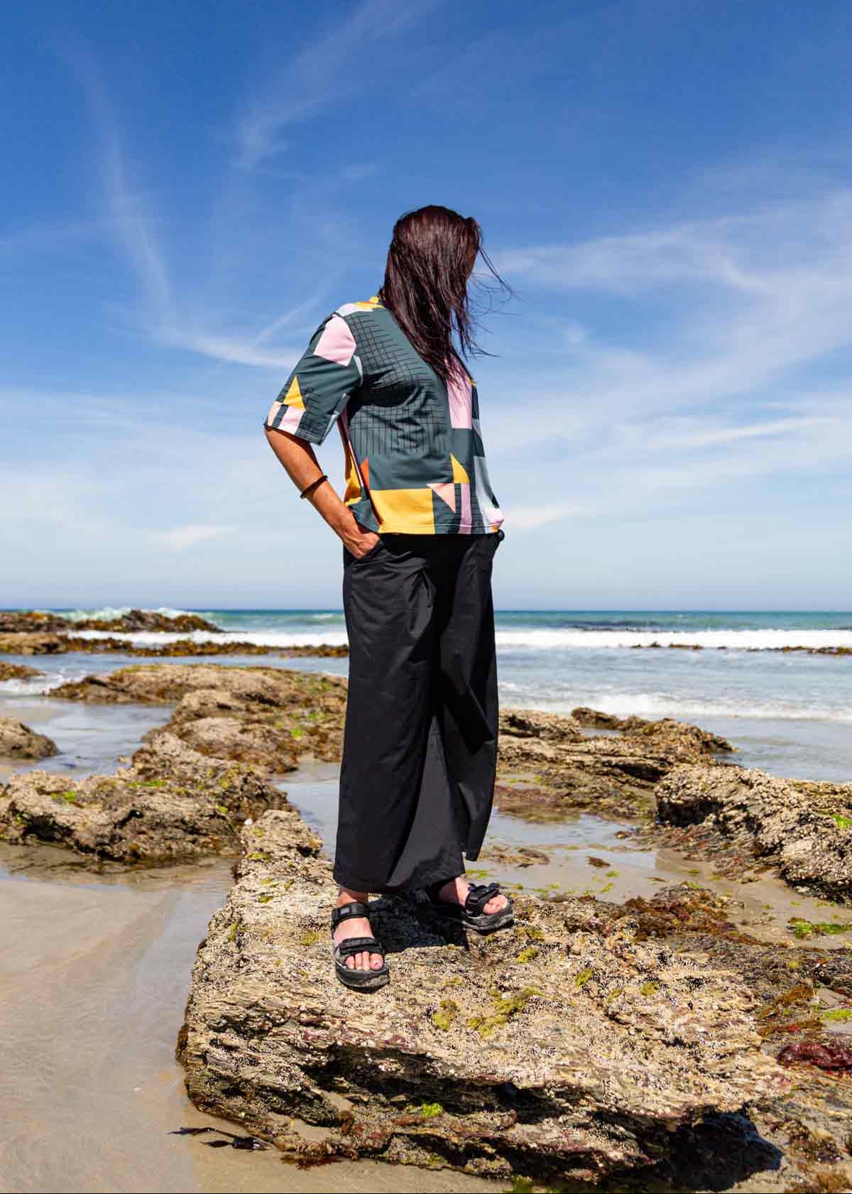 Woman with long dark hair standing on rocks at a beach with her hands in the pockets for the black Asmuss Elsie Wide Leg Trousers. She is also wearing the Asmuss Drew T-shirt in a geometric print