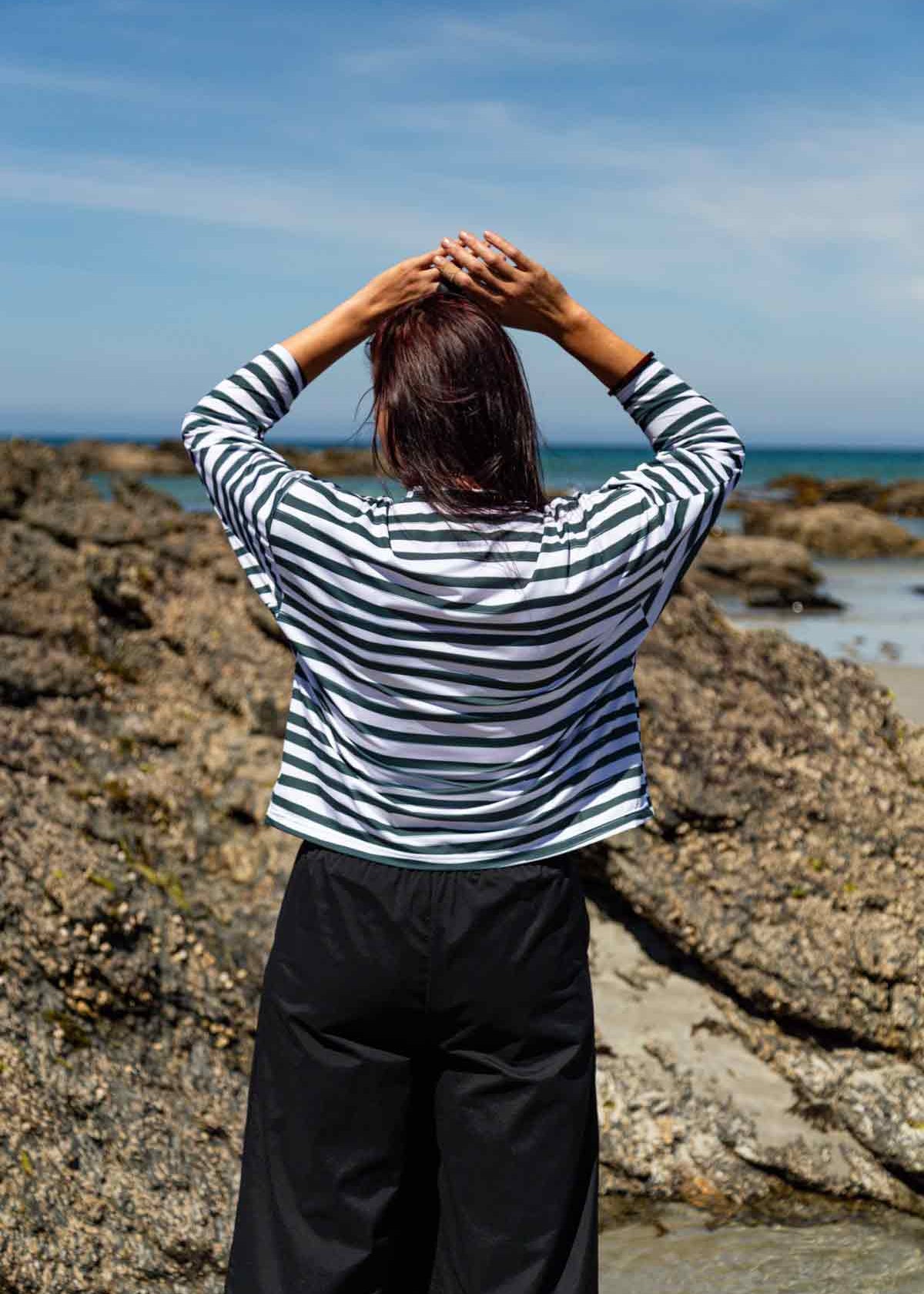 Woman with dark hair facing towards rocks and the blue sea with her hands on her head. She is wearing the Asmuss Zaha Puzzle Top with horizontal painterly stripes in white and dark green.