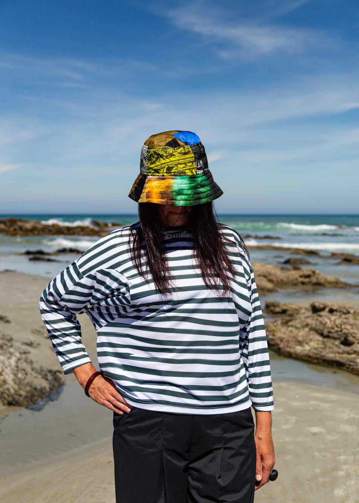 Woman with dark hair standing in front of rocks and the ocean with her right hand on her hip. She is swearing the Asmuss Zaha Puzzle Top with horizontal painterly stripes in white and dark green and a brightly coloured bucket hat