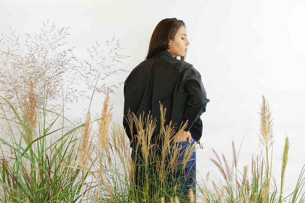 Woman standing with her back to us in amongst tall grasses wearing the Asmuss Roam jacket in black Bio Castor Bean fabric