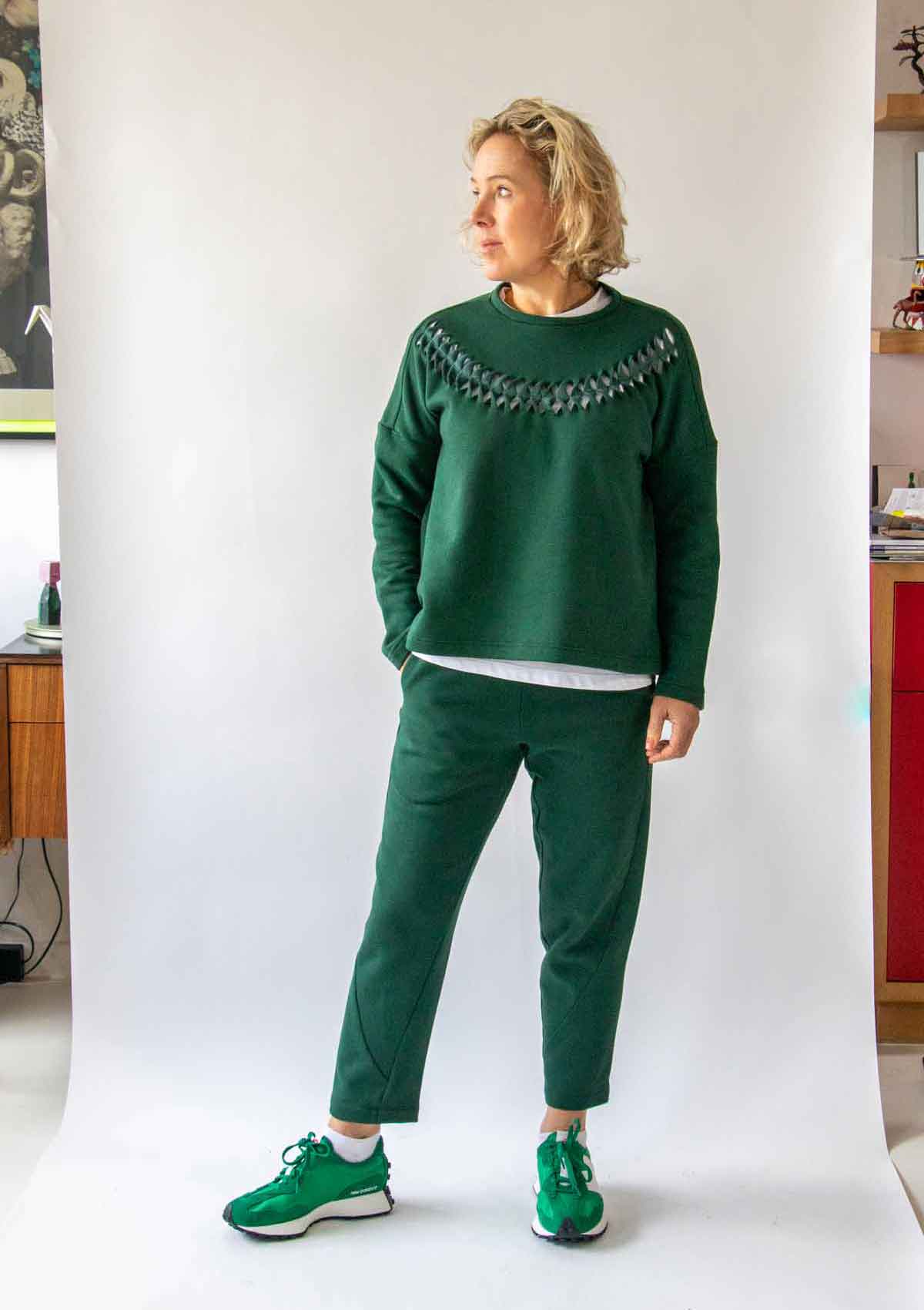 2.	Woman looking to her right, with short wavy blond hair, wearing the Asmuss Anni Sweatshirt in Trekking Green with the Cut Twist and Stitch detailing and the Asmuss Curve Joggers in Trekking Green and green trainers