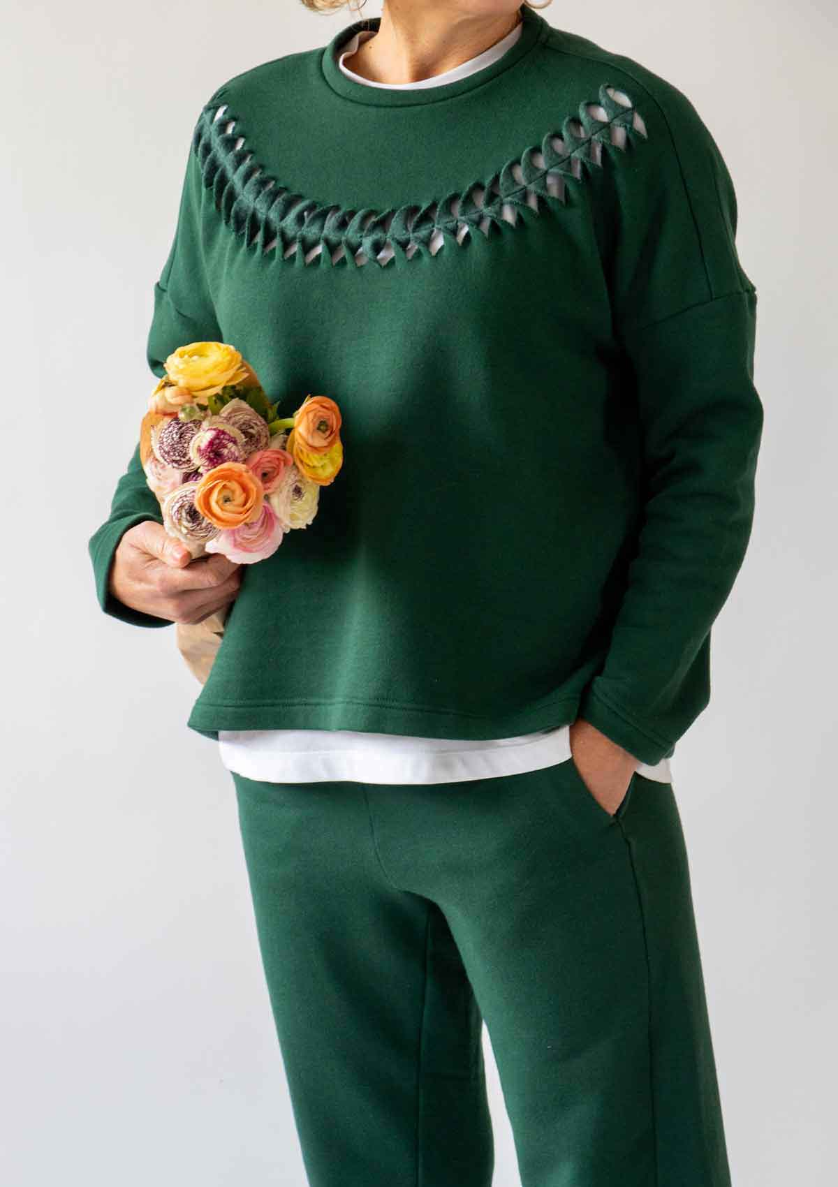 1.	Close up of a woman wearing the Asmuss Anni Sweatshirt in Trekking Green with the Cut Twist and Stitch detailing while holding a bunch of yellow, orange and pink flowers under her arm