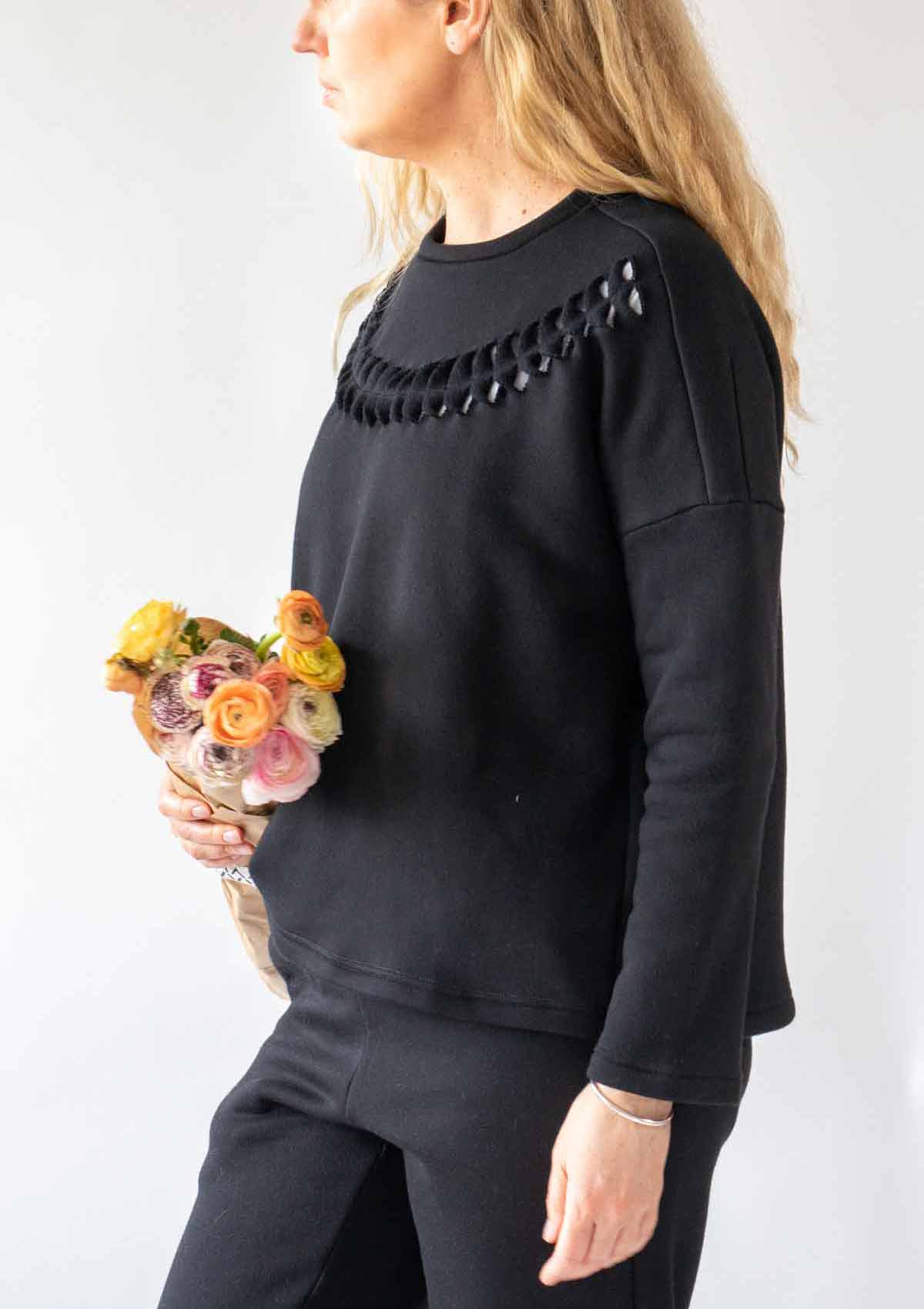 Woman with long wavy blonde hair standing side on wearing the Anni Sweatshirt in Black with cut twist and stick detailing on the front. She is carrying a paper wrapped bunch of flowers in yelllow, pink and orange