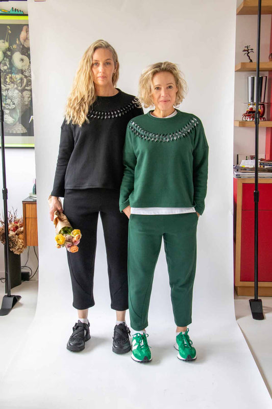 Photo of two women wearing the Asmuss Anni Sweatshirts and Curve Joggers one holding a small paper wrapped bunch of flowers. They are standing in front of a white background but the eclectic shelves of the space can be seen