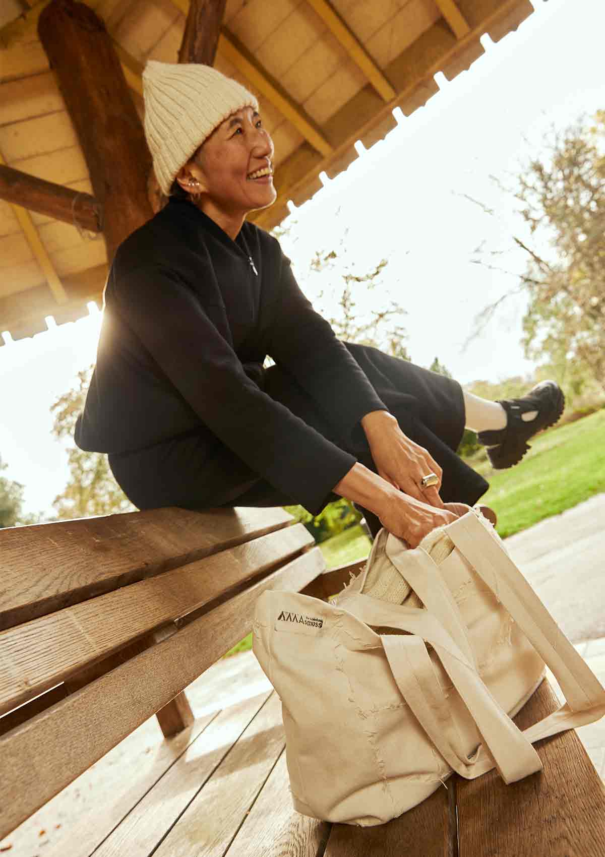 Woman sitting on bench in gazebo, wearing the Asmuss Hepworth zip through sweat, Column joggers in black and also an Asmuss hand knitted ecru beanie. The women is laughing and reaching down into the Asmuss Scraps Tote bag 