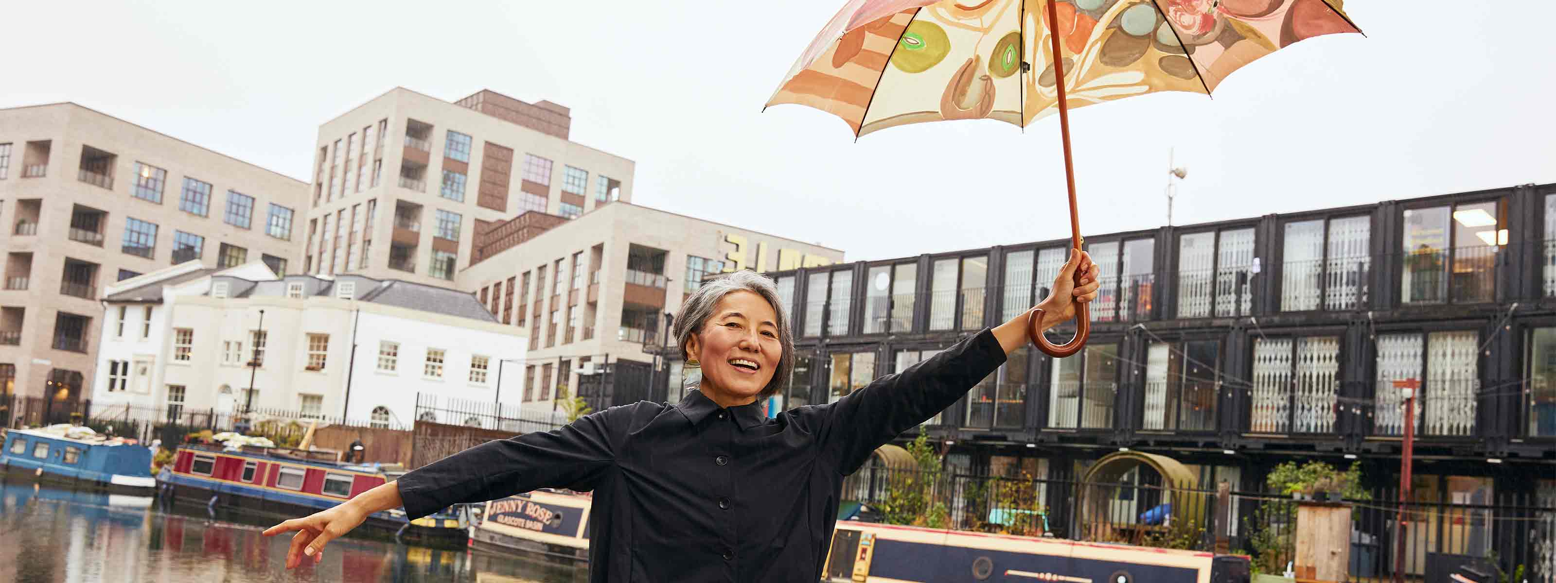 Women smiling holding a colourful umbrella up in the air with one hand and the other hand pointing backwards, it looks like she is enjoying being Mary Poppins. She is wearing the Asmuss Roam Jacket and Eddy Cargo Trousers.  She is standing in front of the Regent Canal in London