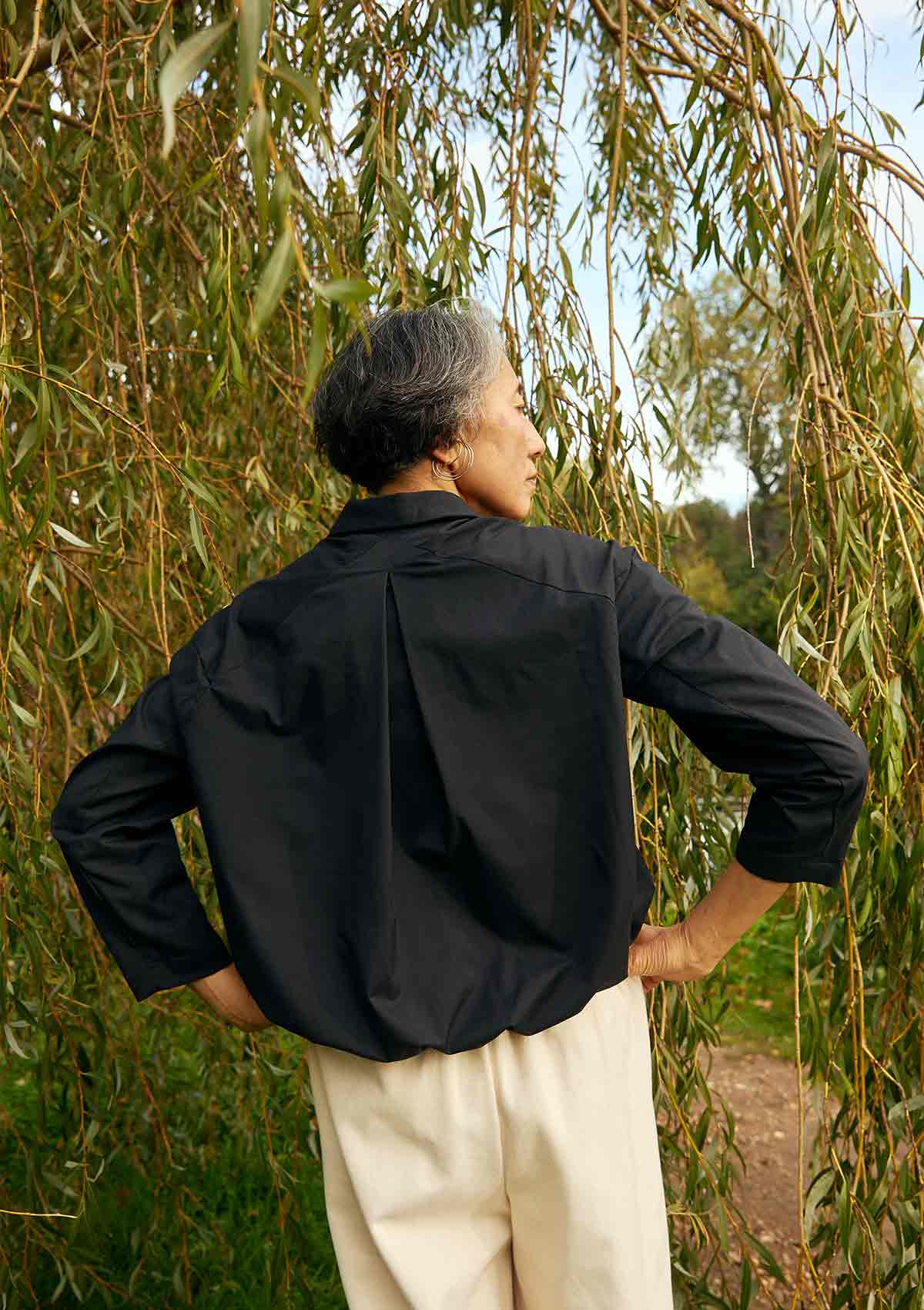 Women, standing back on, wearing the Asmuss Roam Jacket showing the inverted pleat detail on the back, with her hands on her hips. She is standing in front of willows in a park