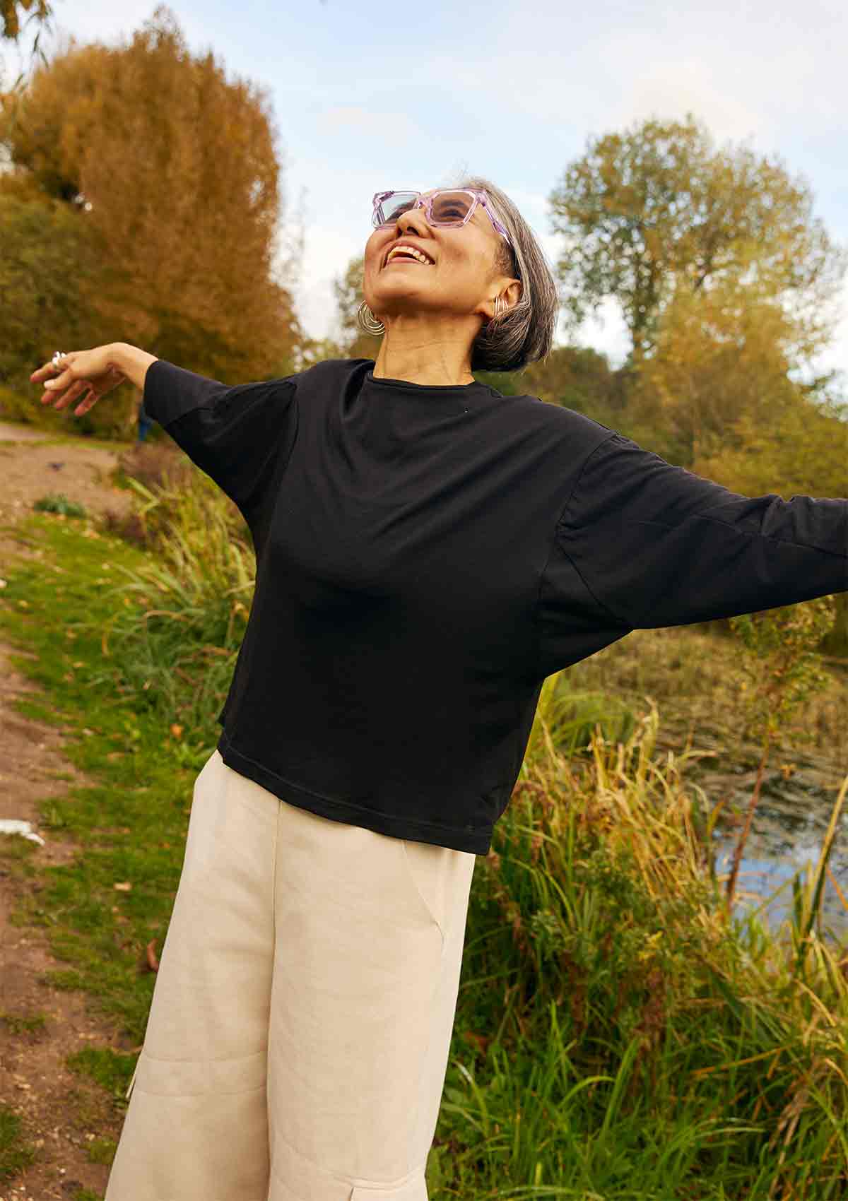 Woman looking up into the sky with her arms out.  You can see the bat wing sleeves of the Asmuss Puzzle Top that she is wearing.  She is standing in a field with trees and pond behind her.
