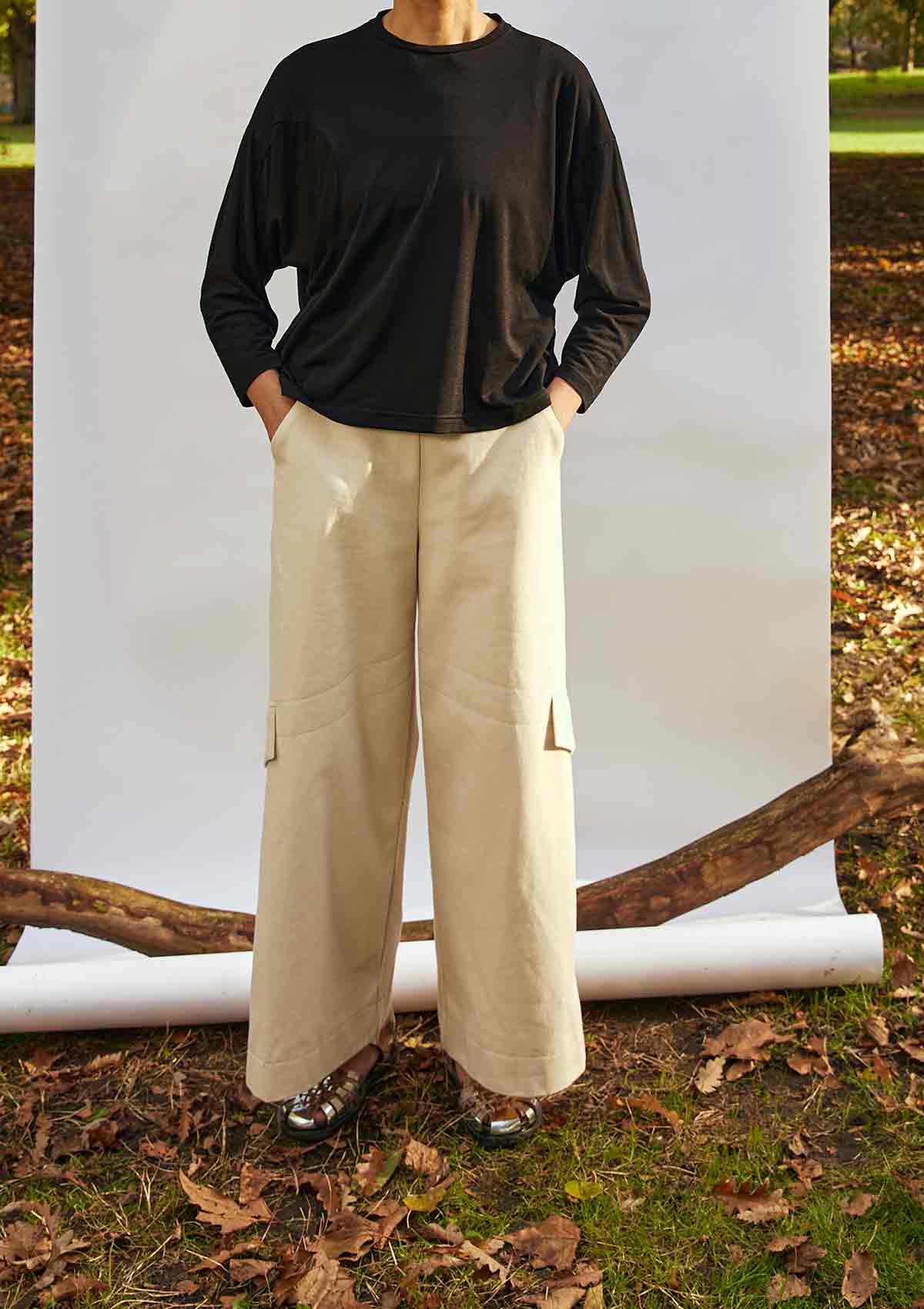 Woman standing face on wearing the Asmuss Eddy Cargo Trousers  and Asmuss Puzzle Top.  The hem buttons are loose.  She is standing in front of a white background, weighted down by a branch in a park