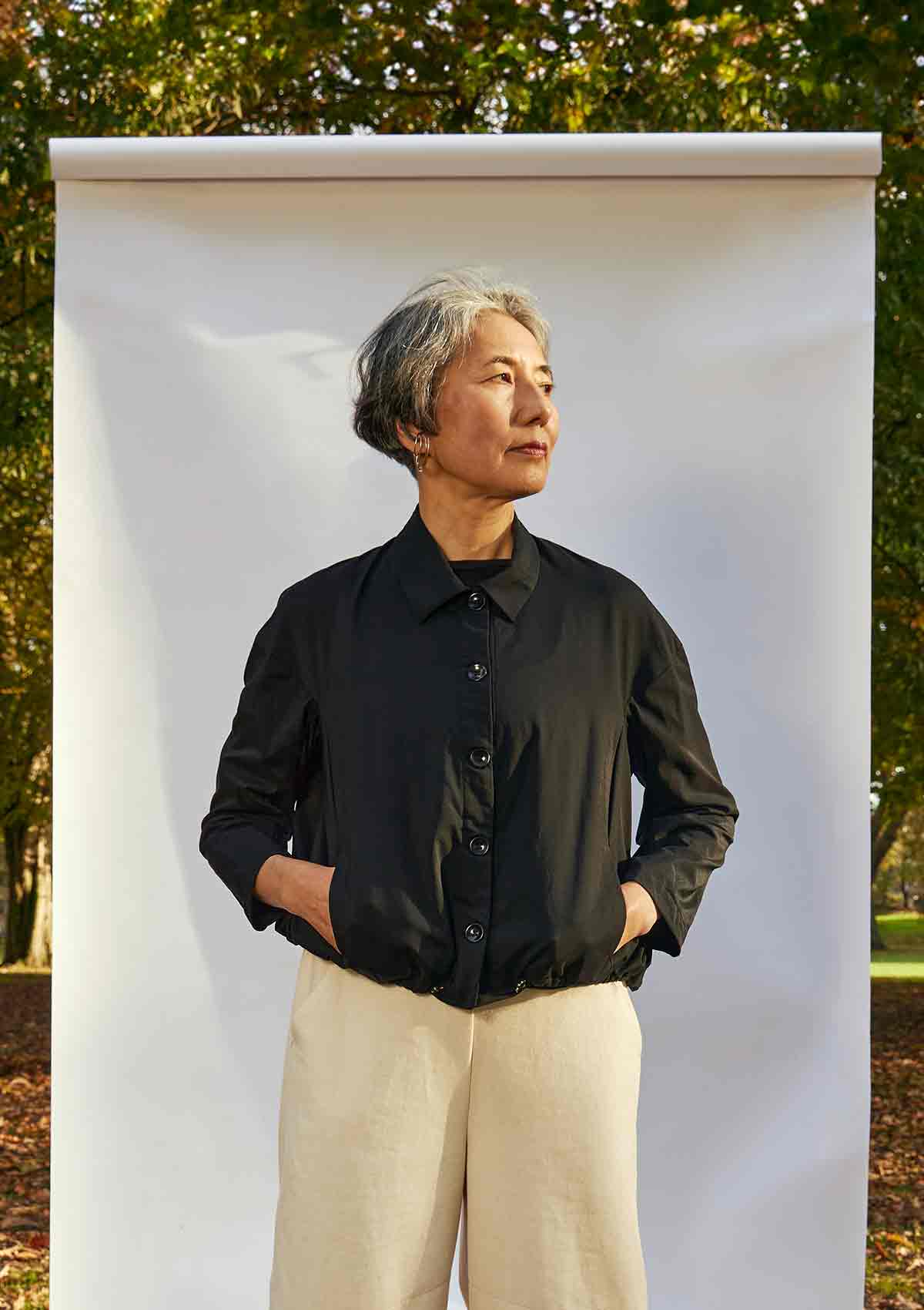 Women, standing front on, wearing the Asmuss Roam Jacket buttoned up with her hands in the side pockets. She is standing in front of a white background in a park