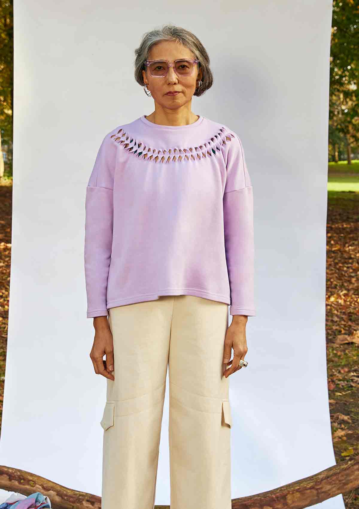 Women standing face on wearing the Anni Sweatshirt in Lilac with detailing where the fabric is cut, twisted and stitched back in place. She is standing in front of a white background in a park