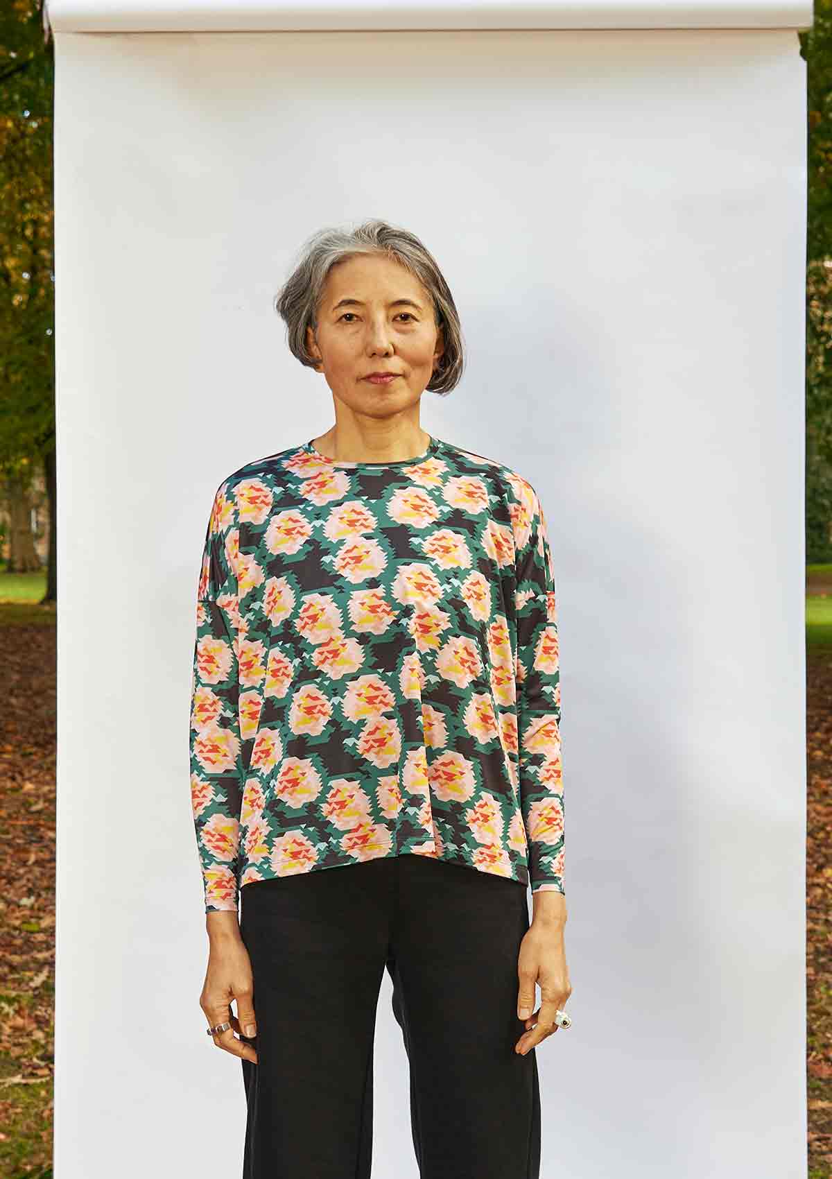Woman standing face on in front of a white background that is outside in a park. She is wearing the Asmuss Paige Long Sleeve T-shirt in a geometric rose print recycled polyester fabric.  The top is untucked so how the fabric falls can be seen