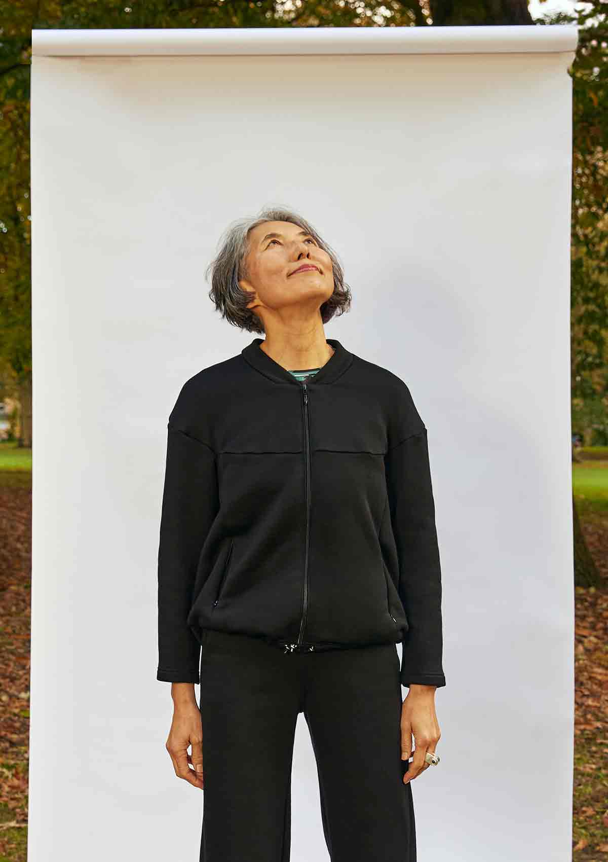 Woman, looking up, standing with her hands by her sides. She is wearing the Asmuss Hepworth Zip Through Sweatshirt zipped up.  She is standing in front of a white background outside in a park 