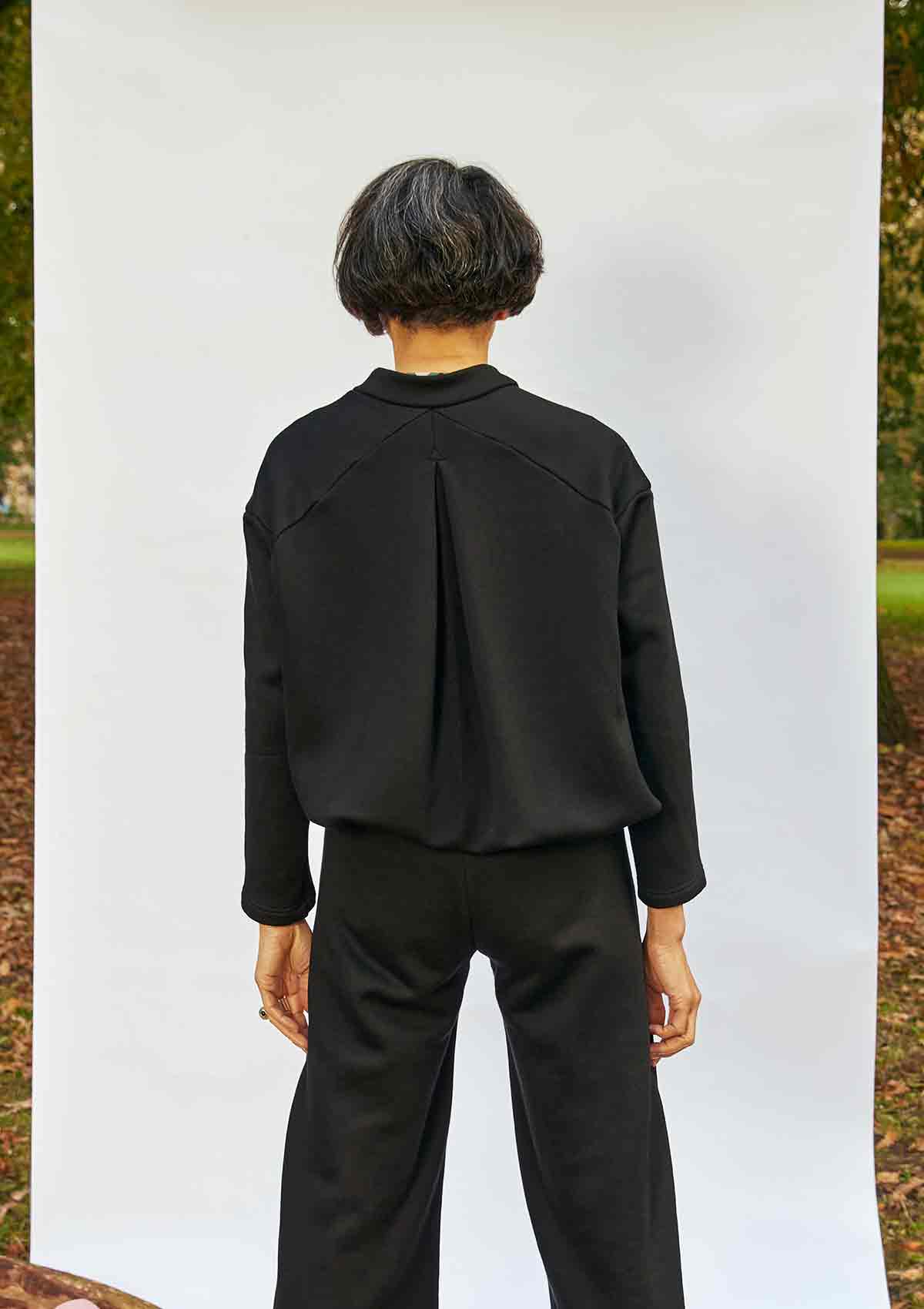 Woman stadning back on showing the inverted pleat detail on the Asmuss Hepworth Zip Through Sweatshirt.  She is standing in front of a white background that is outside in a park