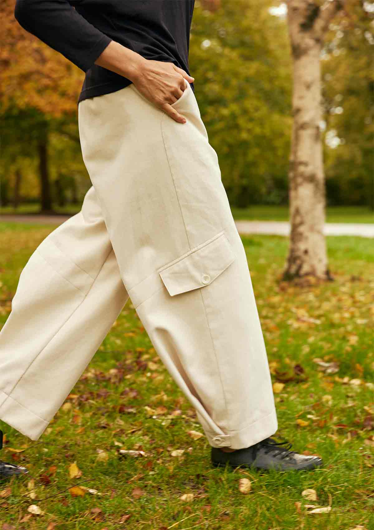 Woman walking in a park with her right leg forward showing the tab pocket detail on the leg and the buttoned hem of the ercu Asmuss Eddy Cargo Trousers.  Her hand is in the hip pocket.