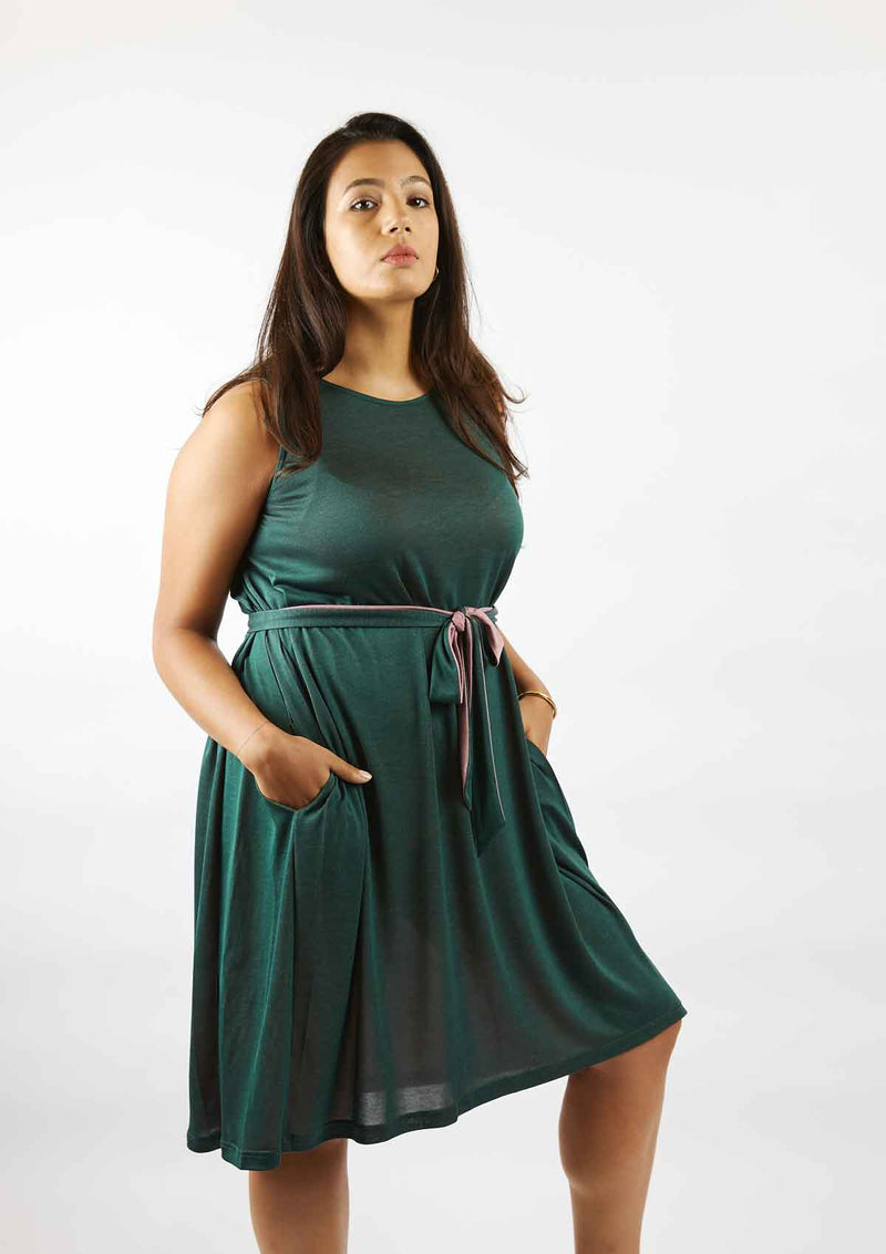Woman standing with her hands of the pockets of the sleeveless pine green Asmuss Drift Dress that has been tied with the self fabric belt