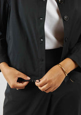 Woman wearing the black Asmuss Roam Jacket showing the drawstring that can be used to create shape in the jacket. The Roam Jacket has a collar, button front and drawstring