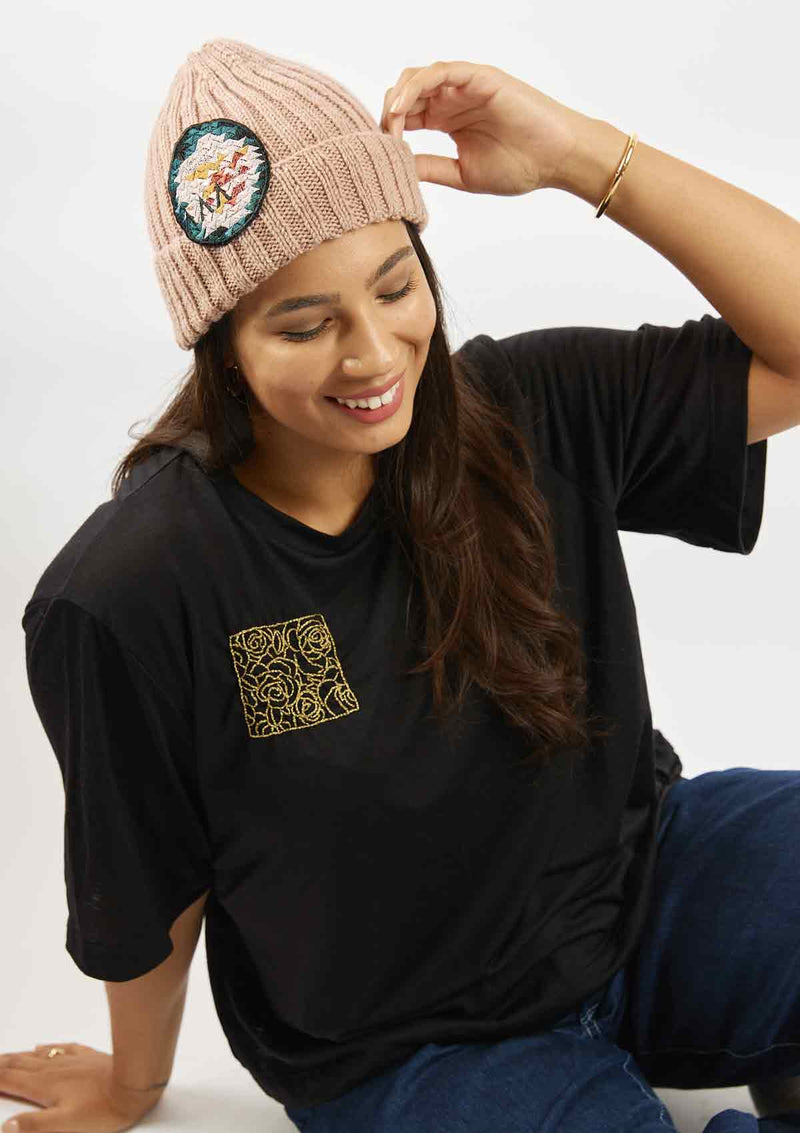 Woman sitting and leaning on her hand wearing the rose embroidery in a square on the upper right of the black Asmuss Evelyne Tshirt that has been hand embellished by Asmuss designer